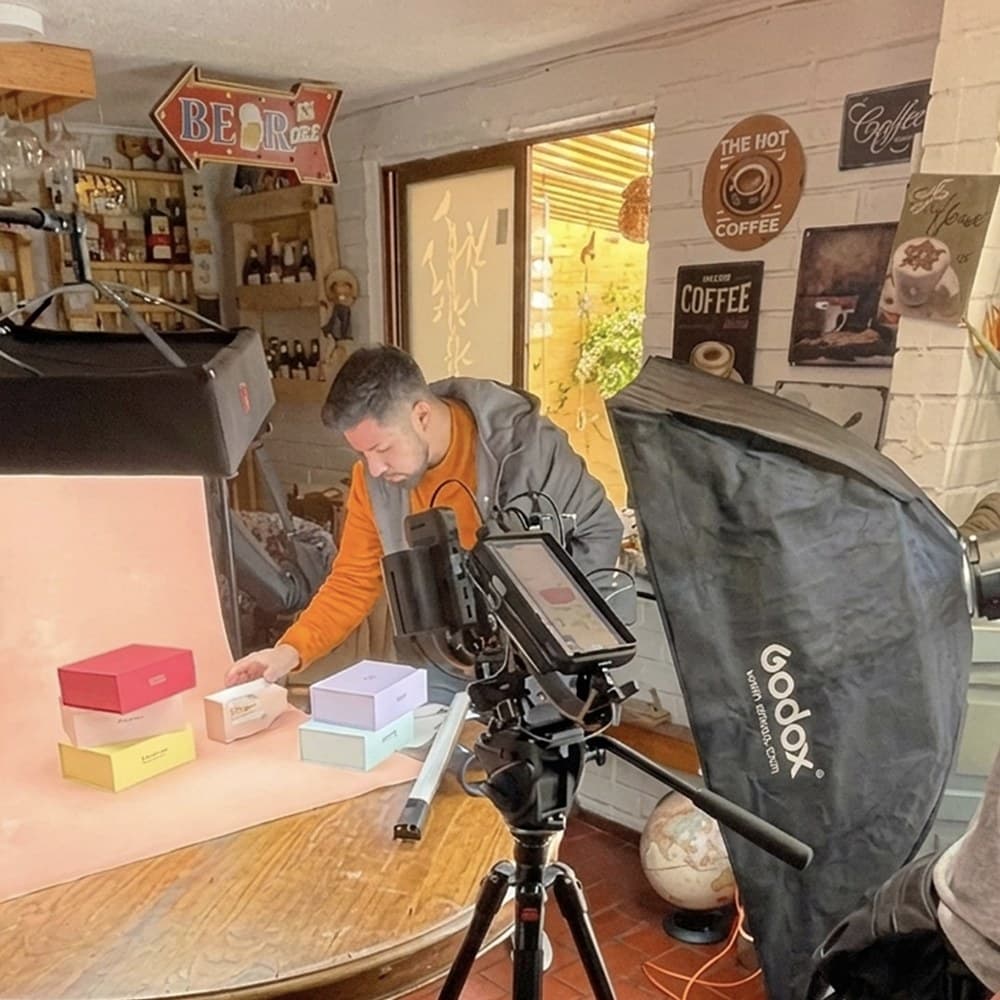 Detrás de cámara de una jornada de TMI para Beppy: una persona ordena cajas de la marca sobre un fondo rosado, frente a una cámara con monitor y una luz con softbox, dentro de un local decorado con letreros de café.